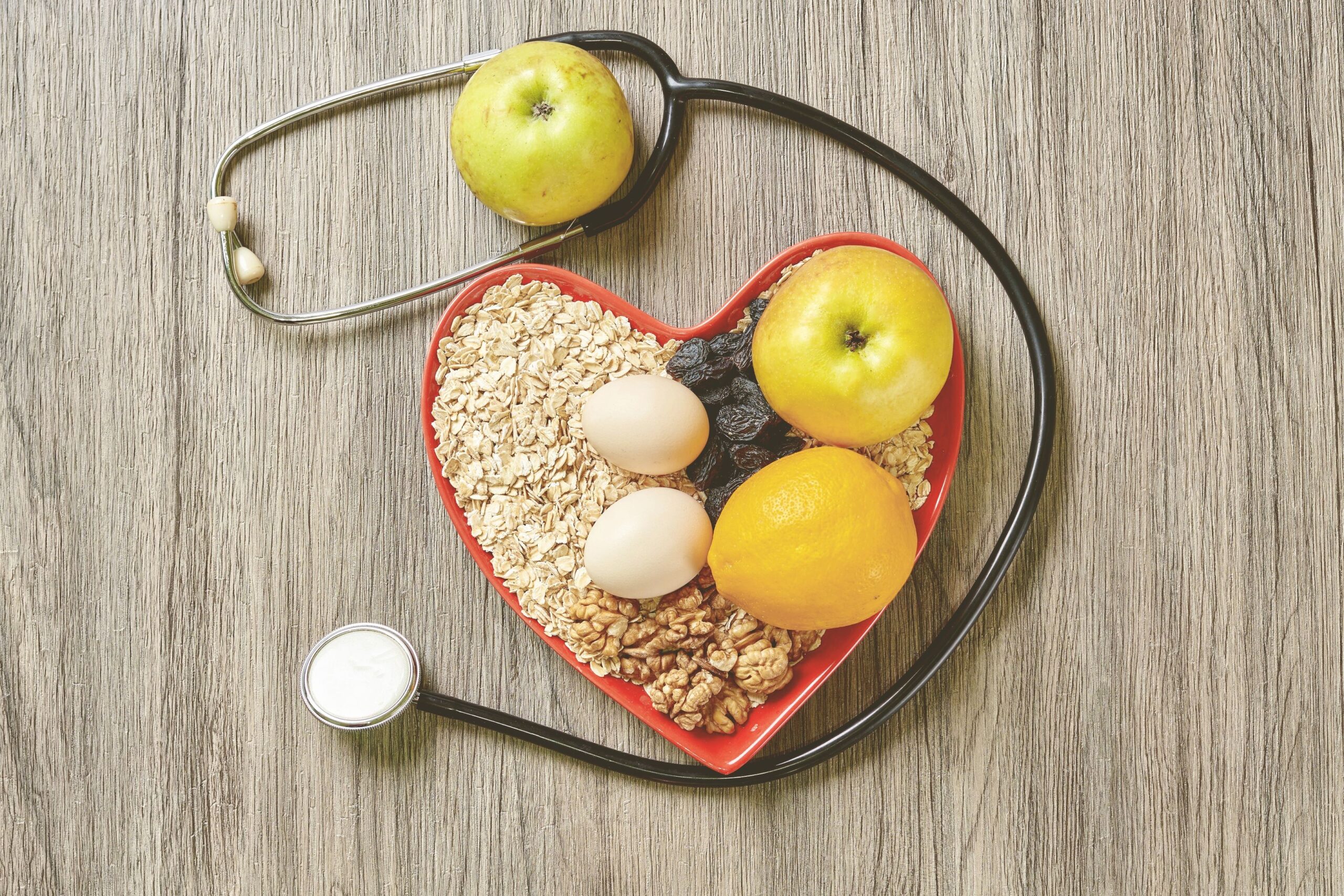 Heart-shaped bowl of healthy foods, including oats, eggs, fruit, and nuts, with a stethoscope nearby.