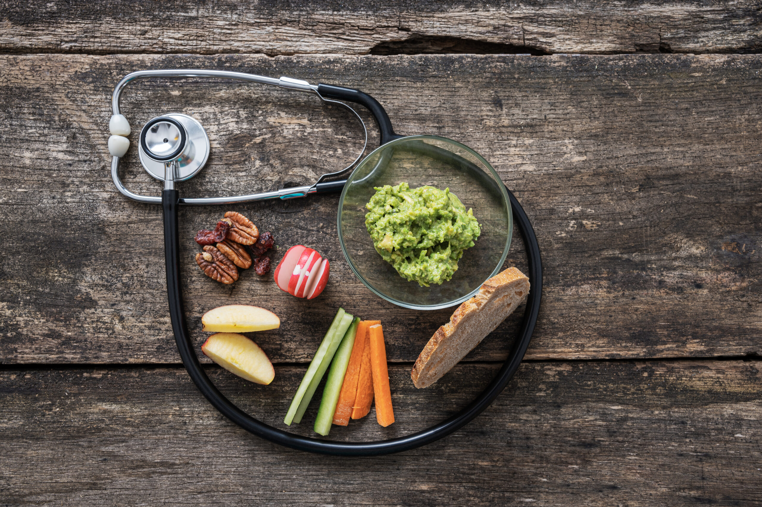 Healthy snack plate with guacamole, fruit, vegetables, nuts, and bread, beside a stethoscope.