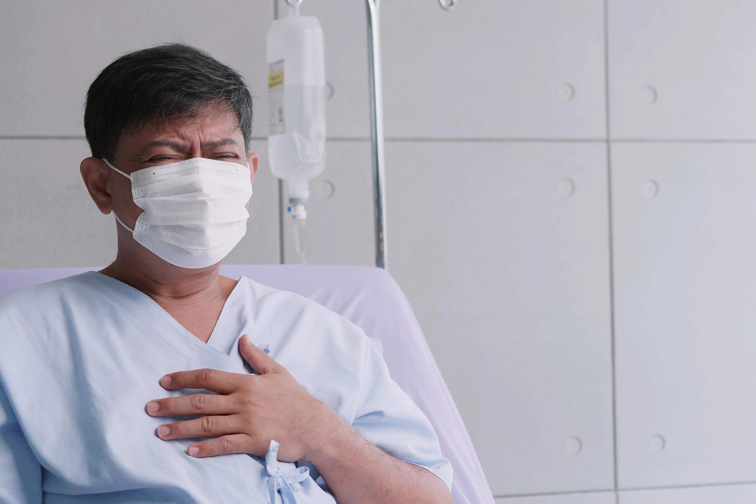 Sick man in hospital bed wearing a face mask, clutching his chest.