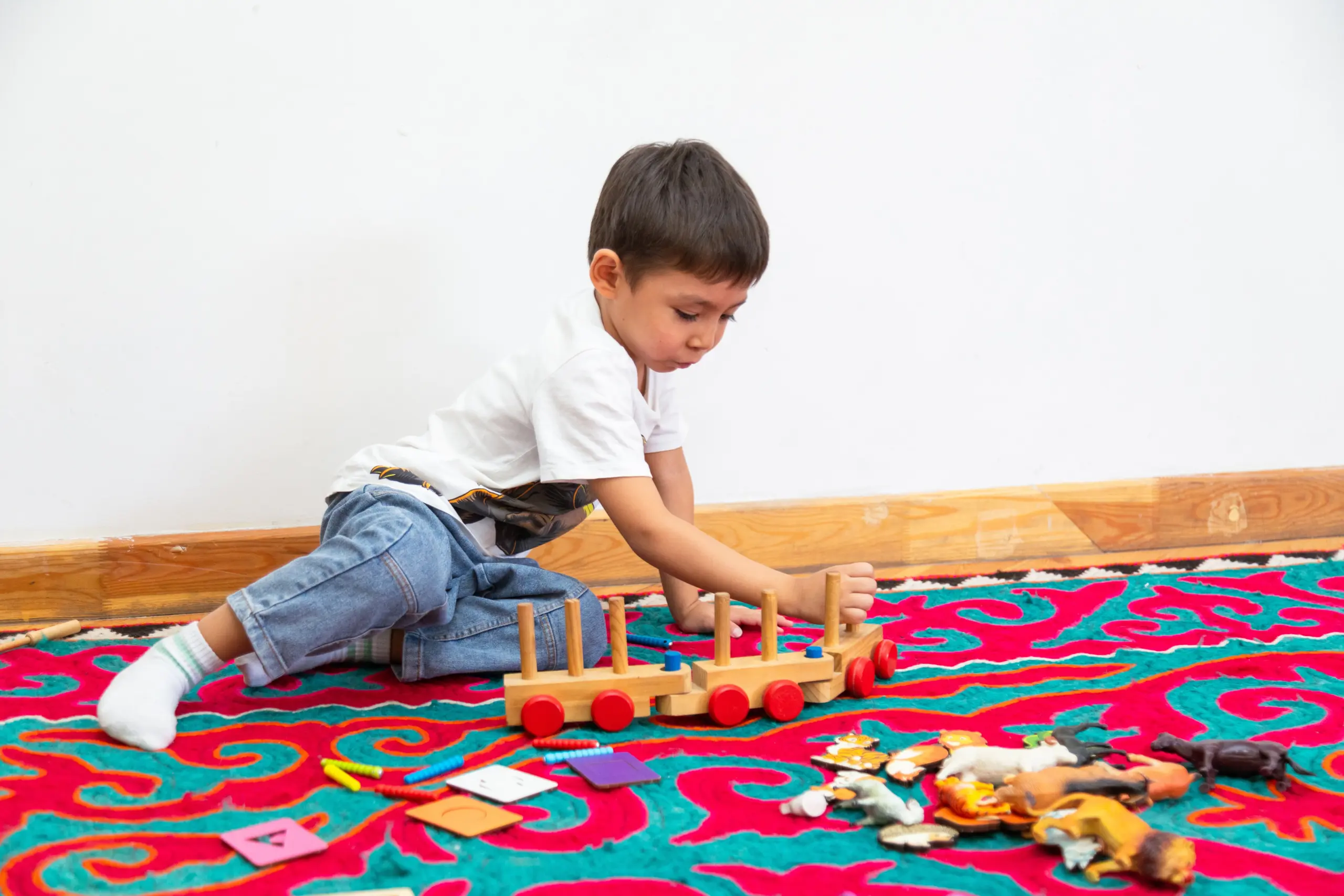 Young boy playing with wooden train and animal toys on a colorful rug.